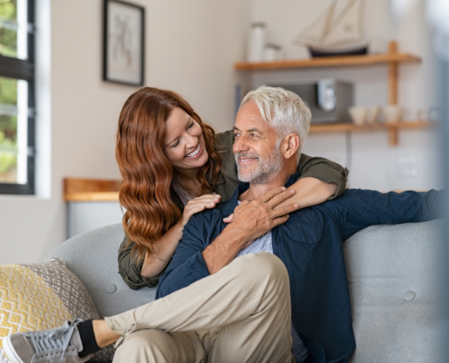 Smiling,Woman,Hugging,Her,Husband,From,Behind,In,The,Living